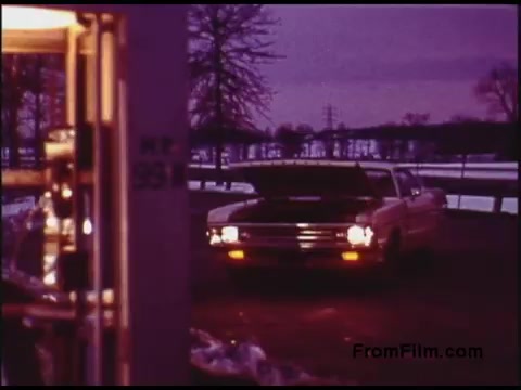 The still depicts a vintage scene from a 1970s Bell System film, showcasing a car parked with its hood open, likely indicating a mechanical issue or maintenance. The backdrop features a dusky sky and bare trees, possibly signaling early evening or winter. The image captures a nostalgic, cinematic quality typical of that era, hinting at themes of technology and everyday life.
