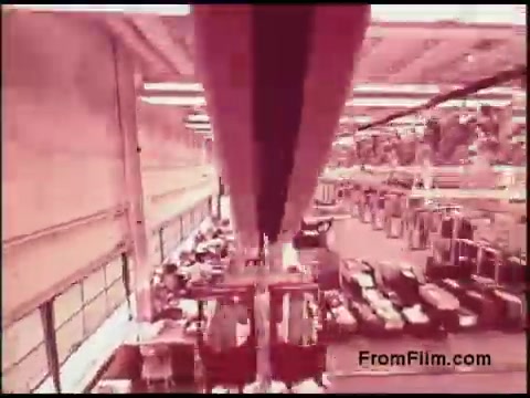 The still from the 1970s Bell System film shows an interior factory or processing facility, with large overhead machinery and a view of workstations below. The color palette has a reddish tint, suggesting a vintage aesthetic common in older films. The space is bustling with activity, indicating a dynamic work environment, possibly related to telecommunications or manufacturing. Overall, it captures a moment of industrial life during that era.