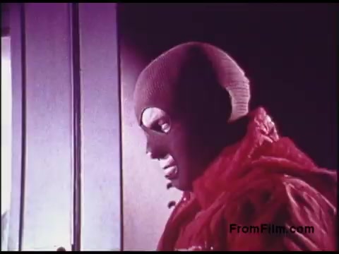 The still from the 1970s Bell System film features a character in a striking costume, complete with a face-covering mask and a bold red outfit. The lighting is dramatic, enhancing the character's expression as they engage with their surroundings. This image evokes a sense of intrigue, possibly relating to themes of communication or security, consistent with the film's context.