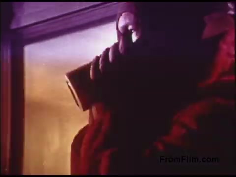 The still from the 1970s Bell System film depicts a figure in a red coat, using a handheld device that resembles a telephone. The background features warm, diffused lighting, hinting at an indoor environment. The scene conveys a sense of communication or monitoring, characteristic of the themes explored in Bell System's promotional or educational films from that era.