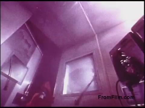 The still from the 'Bell System Film from the 1970s' features a dimly lit interior space, possibly a laboratory or control room. The walls appear somewhat industrial, with various equipment and a window that suggests a view into another area. The color tone is slightly off, giving a retro and muted aesthetic typical of films from that era. A figure can be faintly seen in the lower left, implying some activity or presence in this setting.