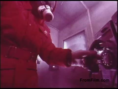 The still depicts a figure in a bright red suit and gas mask, interacting with a telephone in a misty, possibly industrial environment. The atmosphere is tinged with a purple hue, adding to the surreal quality of the image. This scene likely illustrates a safety or communication scenario from a 1970s Bell System film, emphasizing precautions in potentially hazardous conditions.