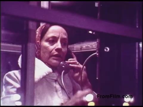 The still from the 1970s Bell System film shows a woman in a phone booth, speaking intently on a telephone. She appears concerned, conveying a sense of urgency or emotion. The color palette and overall aesthetic reflect the era, enhancing the vintage feel. The environment suggests a focus on communication technology and personal interactions typical of that time.