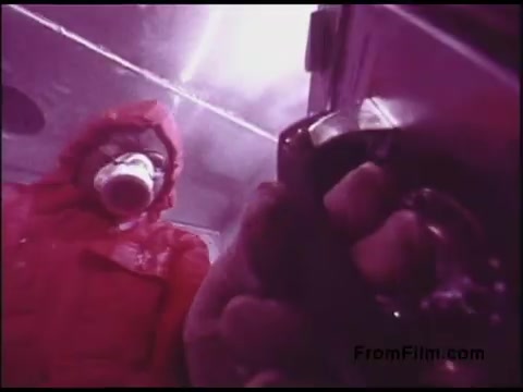 The still features a figure dressed in a bright red jacket with a hood, possibly indicating a cold environment. The individual is holding a cup, suggesting they are taking a break or consuming a hot beverage. In the foreground, a hand is gripping a gadget or instrument, which may imply some technical or operational context. The lighting is a pinkish hue, adding a surreal or stylized quality to the scene. This captures a moment that feels both functional and atmospheric, typical of educational or informational films from that era.