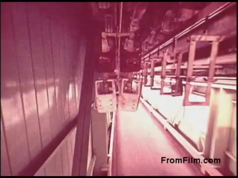 The still features a vintage 16mm film frame from a 1970s Bell System production. It depicts a mechanical or industrial setting, likely showcasing a conveyor system with several suspended carts or pods. The color palette is warm, with reddish hues, indicating the era's film processing style. The perspective suggests a dynamic angle, possibly conveying motion or a unique viewpoint within the facility. Overall, it reflects the technological advancements and aesthetics of the period.