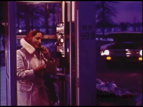 The still from the 1970s film features a woman in a light-colored coat and scarf, engaged in a conversation at a payphone. The scene is set against a twilight backdrop, with a parked car in the background. The atmosphere suggests a candid moment, possibly capturing a nostalgic aspect of communication from that era. The colors are vibrant, likely reflecting the film's vintage aesthetic.