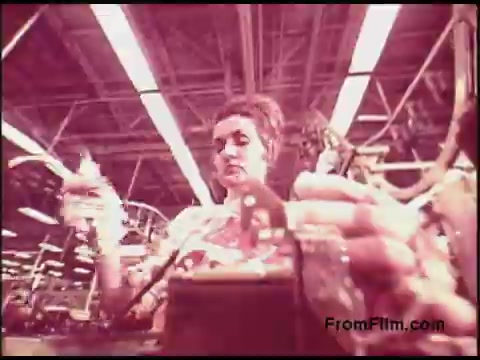The still from the 1970s Bell System film depicts a woman engaged in a manufacturing process, likely at an assembly line. She is holding tools in her hands, surrounded by industrial equipment in a factory setting. The color tone gives a vintage feel, characteristic of the era's film quality. The focus suggests an emphasis on the intricacies of her work within a technological context.