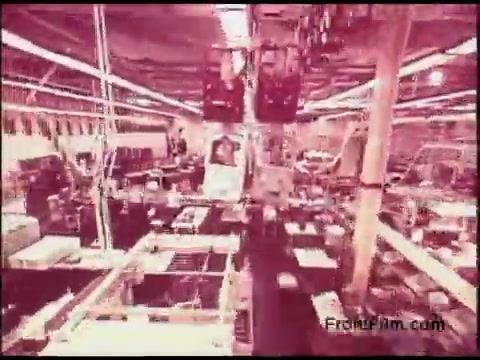 The still from the 1970s Bell System Film depicts a large, industrial workspace filled with various equipment and machinery. Overhead, large structures suggest an organized, potentially automated environment. The lighting casts a warm hue, adding a nostalgic feel, while the overall composition conveys a sense of scale and activity typical of mid-20th century technology-focused settings.