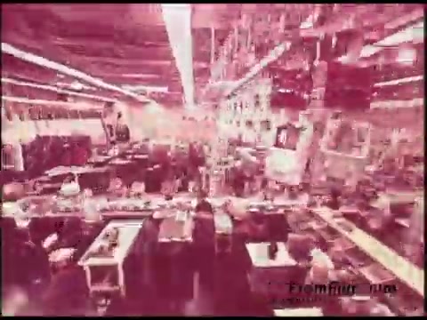 The still from the 16mm film features a bustling factory or manufacturing environment typical of the 1970s. The scene is characterized by numerous workstations filled with equipment and materials. Employees are visible, engaged in various tasks, contributing to a lively production atmosphere. The overall color palette appears reddish, giving it a vintage look that resonates with the era of the film's creation.