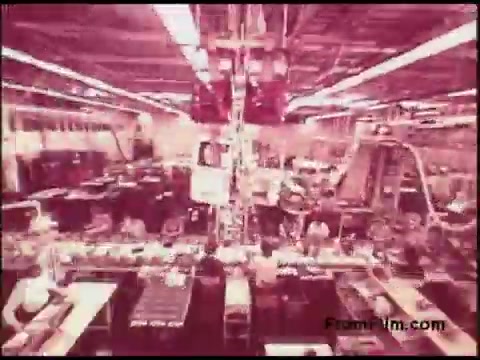 The still from the 16mm film depicts a bustling manufacturing environment, likely from the 1970s. The scene features a large factory with rows of machinery and workers engaged in various tasks. The overhead perspective provides a sense of the scale and organization of the workspace, highlighting the industrial atmosphere typical of that era. The color palette appears muted, characteristic of older film prints, contributing to a vintage feel.