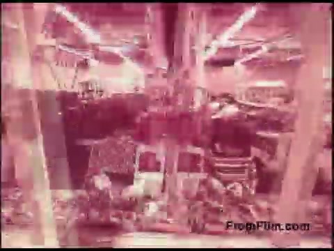 The still from the 'Bell System Film from the 1970s' features a densely packed interior, likely an industrial or telecommunications setting, with various equipment and materials visible. The lighting appears to have a pinkish hue, giving the image a distinct retro aesthetic. The scene suggests a focus on the operational aspects of technology of that era.