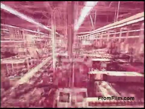 The still features an industrial setting from a 16mm film, likely depicting an interior of a factory or manufacturing space. The image has a reddish hue, suggesting an older film stock or processing effect. Numerous machines and equipment are visible, highlighting a busy production environment typical of the 1970s era. The overall composition gives a sense of scale and activity in the manufacturing process.