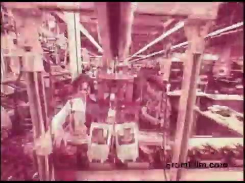 The still features a scene from a 1970s Bell System film, depicting a factory or assembly line environment. The image includes workers engaged in what appears to be the manufacturing or assembly process, surrounded by machinery and production equipment. The pinkish hue suggests a retro aesthetic typical of films from that era. Overall, it captures a snapshot of industrial activity and workforce engagement during that time.