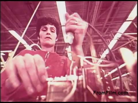The still captures a focused individual working on equipment in a factory setting, likely related to telecommunications or electronics, given the context of the Bell System. The person is engaged in hands-on tasks, with wiring and tools visible, set against a backdrop of industrial lighting and overhead structures. The color tones suggest a distinctive 1970s aesthetic.