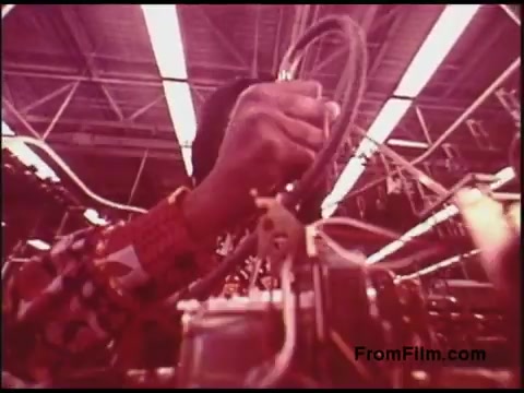 The still from the 'Bell System Film from the 1970s' depicts a close-up of a hand manipulating cables or wires in a factory setting. The individual is wearing a patterned shirt, and the environment features visible overhead lighting, suggesting an industrial atmosphere focused on telecommunications equipment assembly or maintenance. The colors exhibit a distinct vintage quality typical of 16mm film from that era.