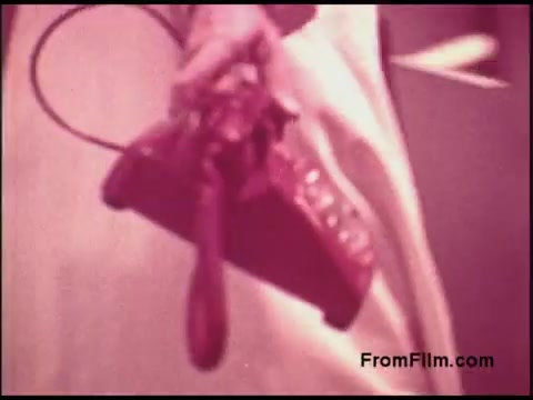 The still from the 16mm film features a hand holding a vintage red telephone, which is likely a rotary or an early push-button model. The background appears somewhat blurred, giving a sense of depth, and the overall color tone leans towards a warm, nostalgic palette characteristic of films from the 1970s. This image captures a moment that reflects the design and technology of that era, possibly emphasizing communication themes related to the Bell System.