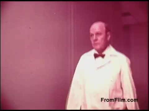 The still features a man dressed in a white coat and bow tie, walking through a corridor. The image has a soft focus, characteristic of 16mm film from the 1970s, and exhibits a warm, faded color palette. The context suggests it is part of an educational or promotional film related to the Bell System.