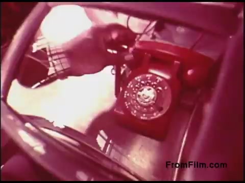 The still features a close-up view of a classic red rotary telephone, likely from the 1970s, positioned on a surface with a hand reaching toward it. The image captures a nostalgic perspective, reflecting the era's iconic design and technology. The colors are vivid, typical of film processing from that time.