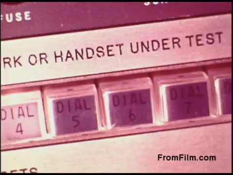 The still features an old telephone testing device from the Bell System in the 1970s. It prominently displays a control panel with labeled buttons for 'DIAL 4,' 'DIAL 5,' 'DIAL 6,' and 'DIAL 7,' as well as an instruction indicating 'MARK OR HANDSET UNDER TEST.' The design reflects the technological aesthetics of the era, showcasing the materials and design elements used in telecommunications equipment at the time.