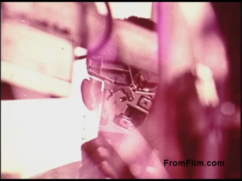 The still from the 'Bell System Film from the 1970s' appears to show a close-up of a piece of equipment or machinery, possibly related to telecommunications. The image is characterized by a warm color palette, dominated by pink and other muted tones, reflecting the aesthetic of the era. There are hints of reflection and depth, suggesting multiple layers or angles in the shot. The context likely revolves around the technology and innovations of that time, possibly illustrating processes or tools used within the Bell System.