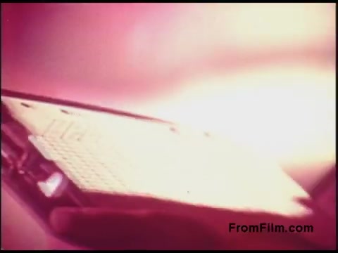 The still from the 16mm film 'Bell System Film from the 1970s' features a close-up of a hand holding an electronic device, possibly a keyboard or small panel. The image is illuminated with a soft, glowing light that gives it a vintage aesthetic characteristic of that era. The background has an abstract, gradient effect, enhancing the retro vibe of the scene.