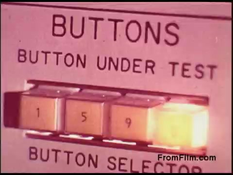 The still depicts a close-up of a testing panel from the 1970s, labeled 'BUTTONS.' It shows a series of buttons with the numbers '1,' '5,' and '9,' and an illuminated button indicating which one is currently under test. The design is utilitarian, reflecting the technological aesthetics of the era, with clear labels and a simple interface. This type of equipment likely relates to telecommunications or electronics testing, emblematic of the innovation in the Bell System during that period.