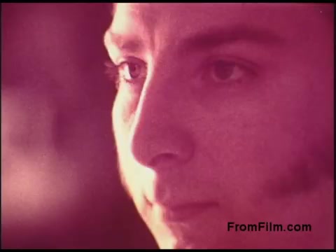 The still is a close-up shot of a young man's face with a soft focus and a reddish hue, typical of film from the 1970s. His expression appears contemplative, showcasing detailed facial features and subtle lighting that emphasizes his eyes. The overall aesthetic is reminiscent of that era's cinematic style, capturing a moment of introspection or emotion.