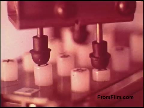 The still from the 1970s Bell System film depicts a close-up of machinery, possibly related to telecommunications or data processing. The focus is on two black components positioned above small white cylindrical objects, suggesting a precise mechanical operation. The lighting creates a warm hue, indicative of the era's film quality. Overall, it conveys a sense of nostalgia and highlights the technological processes of that time.