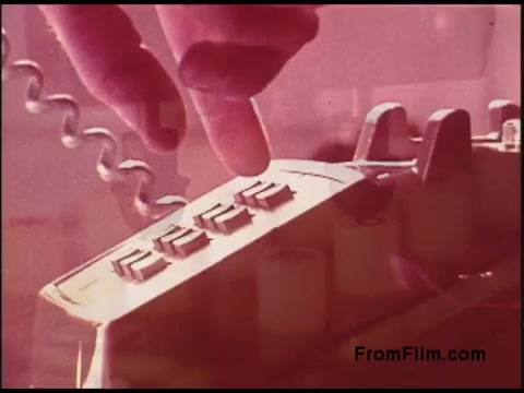 The still features a close-up of a hand poised above a vintage telephone keypad, indicating a dialed number. The image's coloring has a reddish tint, typical of 1970s film stock. The aesthetic reflects the technology of the era, emphasizing telecommunication. The focus on the hand and keypad suggests the critical role of personal communication during that time.