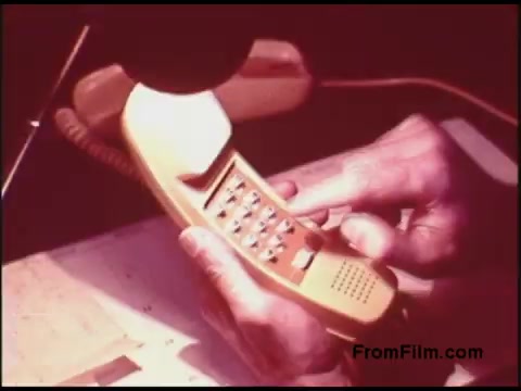 The still depicts a close-up of a person’s hands operating a vintage yellow telephone with a keypad. The image suggests a focus on communication technology from the 1970s, highlighting the design and functionality of telephones during that era. The background appears lightly colored, contributing to a retro aesthetic typical of that time.