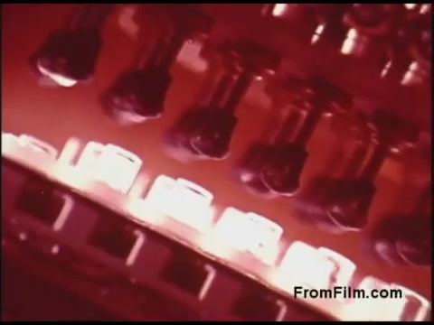 The still appears to be a close-up view of electronic components, showcasing various black sockets or connectors against a backdrop of illuminated elements. The red tint suggests a vintage aesthetic characteristic of the 1970s. This image likely highlights the technology and infrastructure used by the Bell System during that era, emphasizing intricate details.