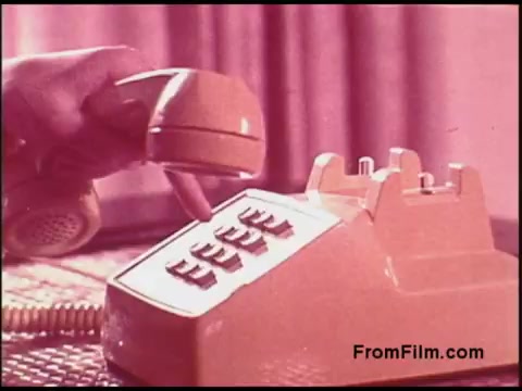 The still features a close-up of a retro telephone from the 1970s, characterized by its bright orange color. A hand is poised to press one of the buttons on the phone, which appears to have a unique design with a horizontal keypad. The background has a soft pink hue, contributing to the vintage aesthetic of the scene. This image is typical of the era and reflects the technology of communication during that time.