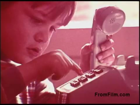 The still features a young boy intently pressing buttons on a classic rotary telephone. The film captures a warm, vintage aesthetic characteristic of the 1970s, with a slightly faded color palette. The setting appears bright and cozy, suggesting a domestic environment. The boy's expression conveys curiosity and focus, reflecting the era's fascination with communication technology.