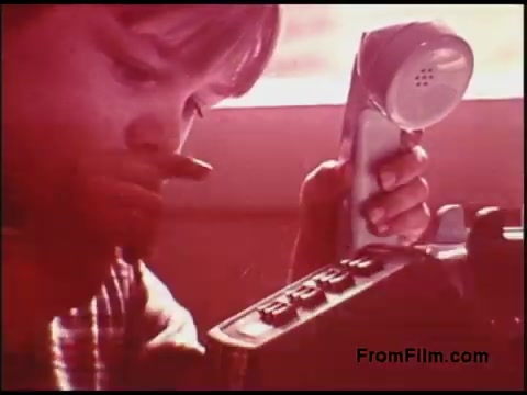 The still from the 1970s Bell System film shows a child closely examining a rotary dial telephone while holding the receiver. The image has a warm, nostalgic color palette, suggesting a focus on communication and technology of that era. The child's expression indicates curiosity or concentration, capturing a moment of interaction with a piece of vintage technology.