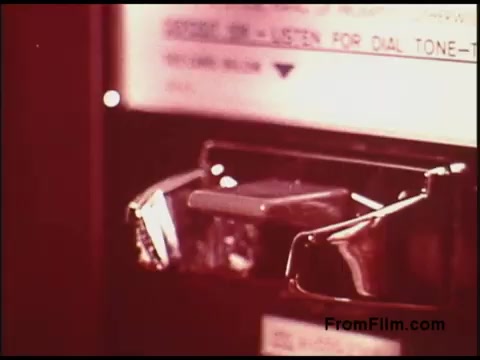 The still features a close-up of a vintage payphone or telephone booth, showcasing the coin slot and dial mechanism. The design reflects the era of the 1970s, emphasizing the technology of the time. Labels and instructions are visible, indicating its use for dialing. The color palette suggests a warm tone typical of films from that decade. Overall, it captures a nostalgic glimpse into communication technology.