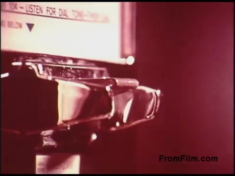 The still from the 1970s Bell System film features a close-up of a payphone, highlighting the receiver and its sturdy design. The background includes instructions for use, emphasizing a time when such communication methods were common. The overall aesthetic is typical of the era, with a warm tint that evokes a sense of nostalgia.