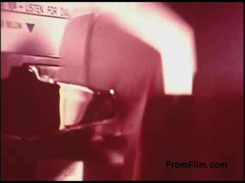 The 16mm still from the 'Bell System Film from the 1970s' shows a close-up of a device, likely a telephone or communication apparatus, with a finger poised to interact with it. The lighting is soft and slightly diffused, giving it a nostalgic quality. The background features a faded sign indicating instructions, perhaps related to dialing or using the device. The overall tone suggests a focus on technology from that era, highlighting both functionality and the design aesthetics of the time.