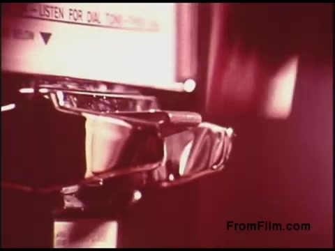 The still features a close-up of a phone dial, likely from a payphone, with a focus on the metallic components and the dial mechanisms. The color tone has a vintage aesthetic, characteristic of films from the 1970s, highlighting the design and technology of that era. The background is softly blurred, drawing attention to the phone.