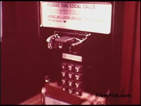 The still features a vintage payphone from the 1970s, showcasing its keypad and a notice instructing users to dial local calls. The design reflects the technology of that era, with a focus on functionality and user guidelines. The overall aesthetic is characteristic of the period, highlighting retro telecommunications equipment.