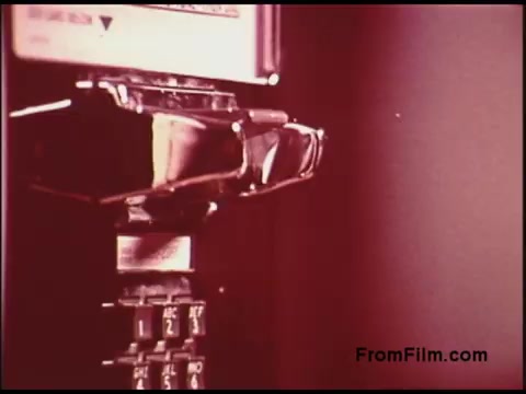The still shows a close-up of a classic payphone from the 1970s. The focus is on the receiver and the keypad, highlighting the metallic finish and the tactile buttons labeled with numbers and letters. The background features a warm, muted color palette typical of the era, giving it a nostalgic feel. The film likely reflects the telecommunications culture of the time, showcasing the payphone as a vital communication tool before the rise of mobile phones.