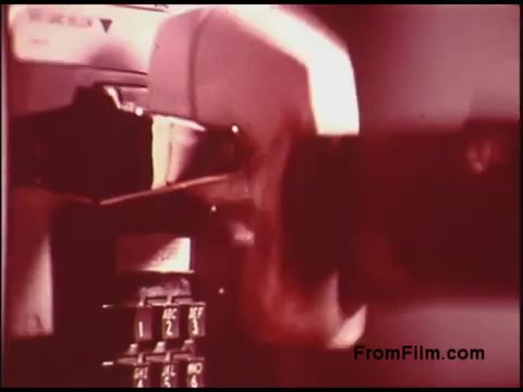 The still from the 16mm film, likely produced by Bell System in the 1970s, features a close-up of a payphone. The image captures a hand inserting a coin into the phone, with the dial pad and phone's structure prominently visible. The color tones suggest a vintage aesthetic, characteristic of films from that era. This snapshot provides a glimpse into the communication technology of the time, showcasing the physical interaction required to make a phone call.