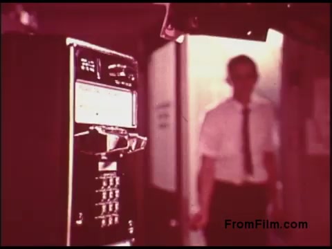 The still features a vintage payphone prominently in the foreground, with a blurred figure of a man in a white shirt and tie walking through a doorway in the background. The film has a warm, reddish tint typical of certain films from the 1970s, suggesting a nostalgic or retro atmosphere. This visual encapsulates a moment in an era when payphones were common, likely reflecting the themes connected to communication and technology of that time.