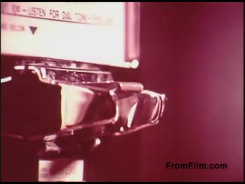 The still features a close-up of an old-fashioned payphone from the 1970s. The payphone's metal casing shines under soft lighting, emphasizing its nostalgic design. Text on the phone suggests it offers instruction for dialing, hinting at its functionality. The overall color tone is warm and slightly muted, characteristic of films from that era, evoking a sense of nostalgia for vintage communication technology.