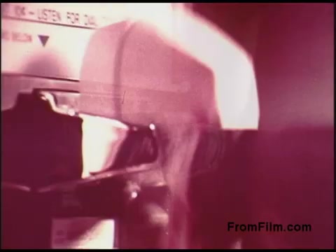 The still from the 1970s 'Bell System Film' features a close-up of a telephone device, possibly a payphone or similar equipment. The image exhibits a warm, slightly faded color palette typical of 16mm film from that era. There are abstract shapes and shadows indicating movement, suggesting someone is interacting with the device. The background contains labels or instructions, hinting at the functionality of the equipment. Overall, the frame captures a moment of communication technology in a historical context.