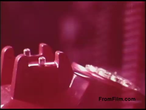 The still features a close-up of a retro red telephone, highlighting its distinctive design from the 1970s. The color saturation is high, giving the image an abstract quality with pinkish hues. The focus is on the phone's dial and receiver, evoking a nostalgic feel associated with the era's communication technology. The background is slightly blurred, suggesting a sense of depth and context without overpowering the main subject.