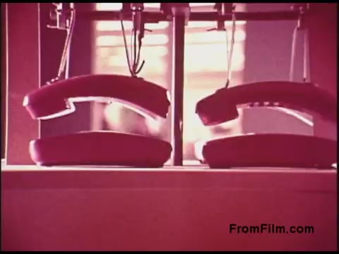 The 16mm still from the 'Bell System Film from the 1970s' features two vintage rotary telephones positioned on a surface. The image has a warm color palette with a slight pink hue, likely indicative of the film's age and processing style. The composition emphasizes the phones, possibly suggesting a focus on communication technology of that era. The background appears softly blurred, directing attention to the central subject.