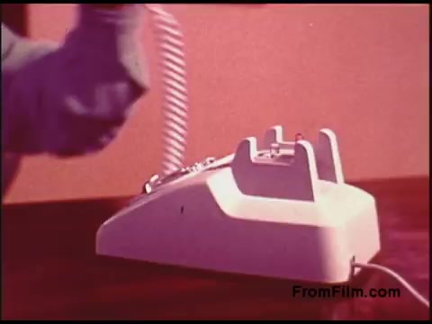 The still features a close-up of a vintage telephone from the 1970s, characterized by a white design and a rotary dial. A hand is seen lifting the receiver, with the background showing a solid color, likely contributing to the film's instructional or promotional context. The overall aesthetic reflects the style and technology of that era within the telecommunication industry.