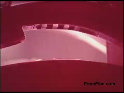 The still features a close-up view of a 16mm film reel, showcasing a portion of the film strip with a prominent red tint. The film appears to be unspooled slightly, revealing a curved section and a contrasting black-and-white striped edge, typical of film leader frames. The overall aesthetic reflects the color processes and graphical style characteristic of the 1970s.