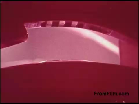The still from the 'Bell System Film from the 1970s' features a 16mm film strip, prominently showing a section of the film and its perforations. The image has a reddish tint, creating a striking visual effect, and suggests an industrial or technical context, likely related to telecommunications. The background is blurred, which keeps the focus on the film itself. This aesthetic evokes a sense of nostalgia for vintage film production techniques.