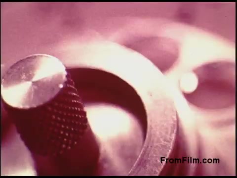 The still from the 1970s Bell System film features a close-up of a mechanical part, possibly from a film projector or other equipment. The image has a soft, warm color palette with a slight pinkish hue, highlighting the textures of the metal. The focus is on a knurled knob and an adjacent cylindrical component, creating a sense of depth through selective sharpness. The composition emphasizes intricate details of the machinery, suggesting a theme related to technology or engineering.